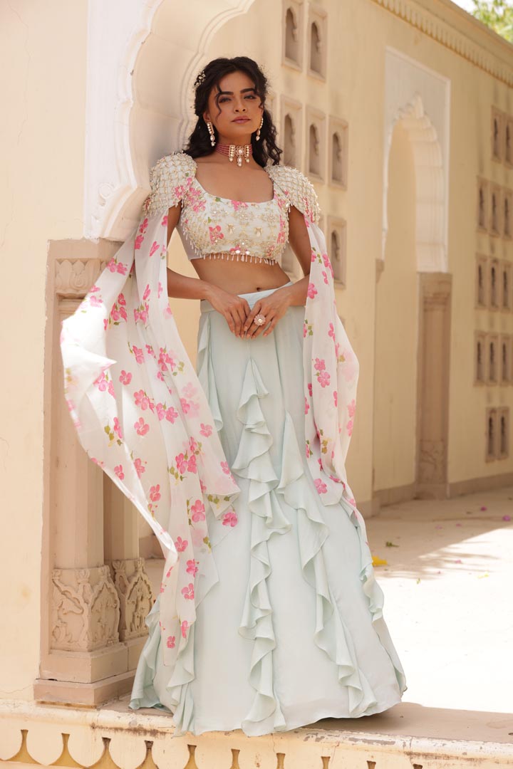 Image of Featuring Georgette Fish cut skirt with ruffle details. Paired with embroidered Chiffon printed blouse, patch of net on both the sides. Cones along with shoulder embroidered cape with floral chiffon flare.