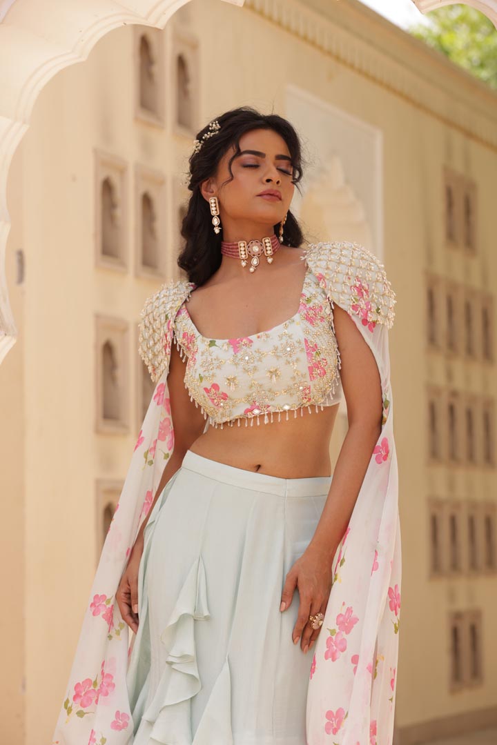 Image of Featuring Georgette Fish cut skirt with ruffle details. Paired with embroidered Chiffon printed blouse, patch of net on both the sides. Cones along with shoulder embroidered cape with floral chiffon flare.
