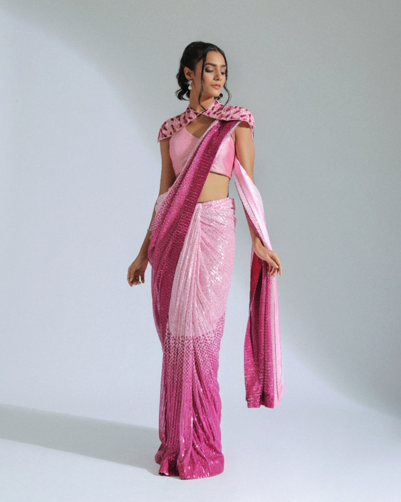 A georgette ombré saree transitioning from light to vibrant shades of Fuchsia pink with sequin lines, accompanied by a cut silk blouse and a removable embroidered shoulder cape. 
