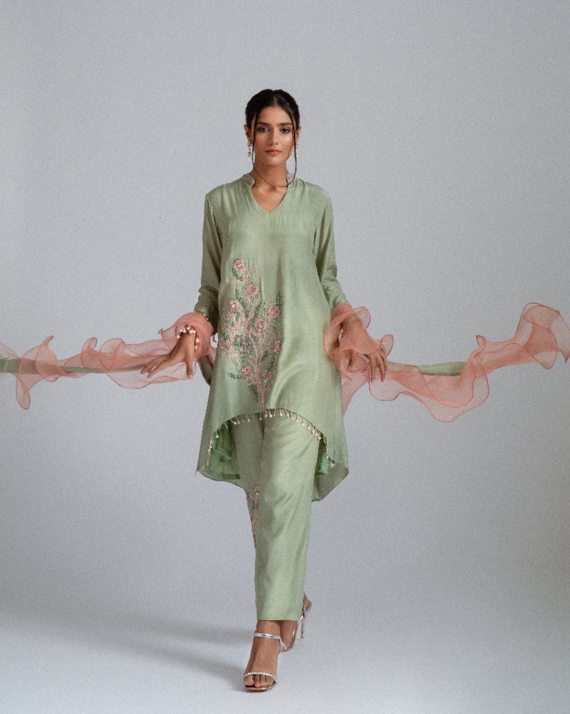 A sage green Silk mul mul tunic shirt with unique back box pleat, embellished with floral motifs, complemented by motif trousers and a peach organza ruffle scarf. 