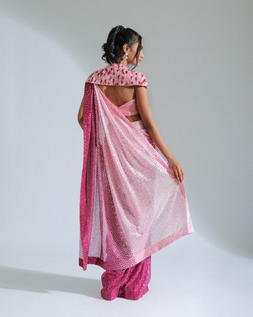 A georgette ombré saree transitioning from light to vibrant shades of Fuchsia pink with sequin lines, accompanied by a cut silk blouse and a removable embroidered shoulder cape. 