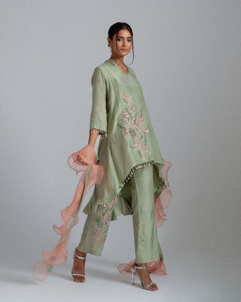A sage green Silk mul mul tunic shirt with unique back box pleat, embellished with floral motifs, complemented by motif trousers and a peach organza ruffle scarf. 
