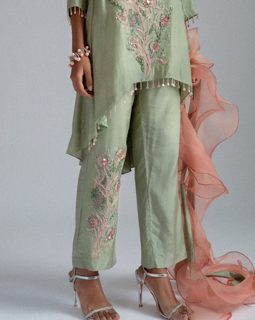A sage green Silk mul mul tunic shirt with unique back box pleat, embellished with floral motifs, complemented by motif trousers and a peach organza ruffle scarf. 