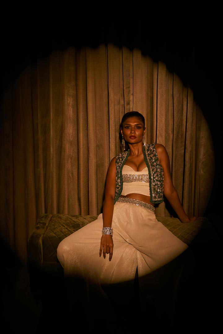 An ivory crushed fabric set featuring flared pants, a crystal-detailed bustier, and an embellished green jacket.