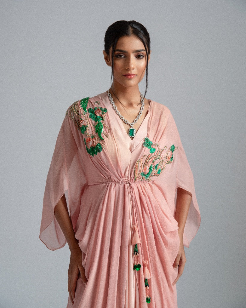 A Peach asymmetrical drape dress paired with a kaftan jacket featuring intricate green and champagne-toned floral embroidery and accentuated by a pleated box opening inner drape. 