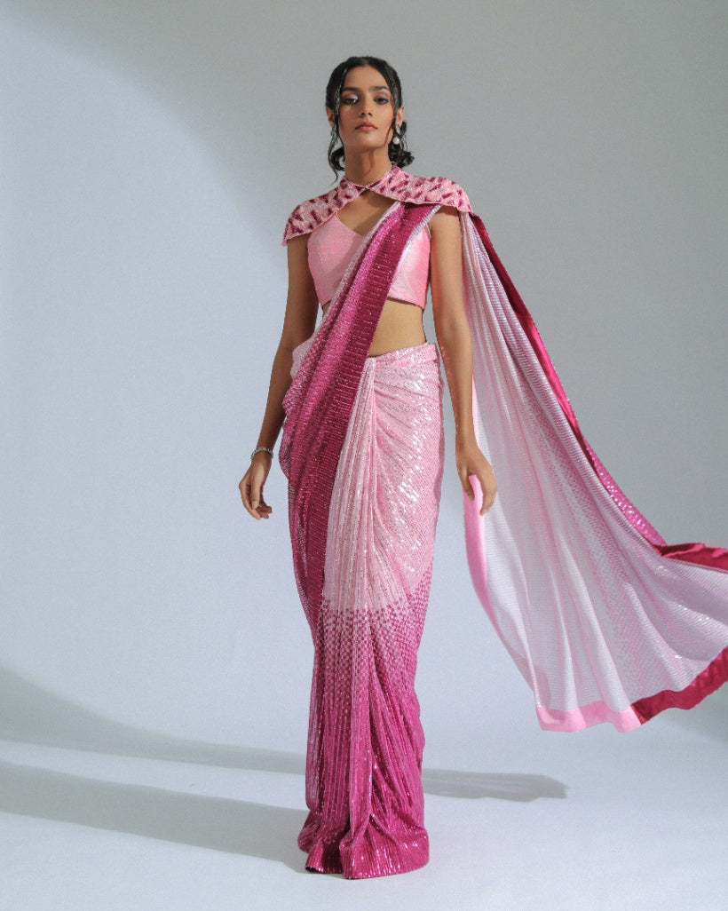 A georgette ombré saree transitioning from light to vibrant shades of Fuchsia pink with sequin lines, accompanied by a cut silk blouse and a removable embroidered shoulder cape. 